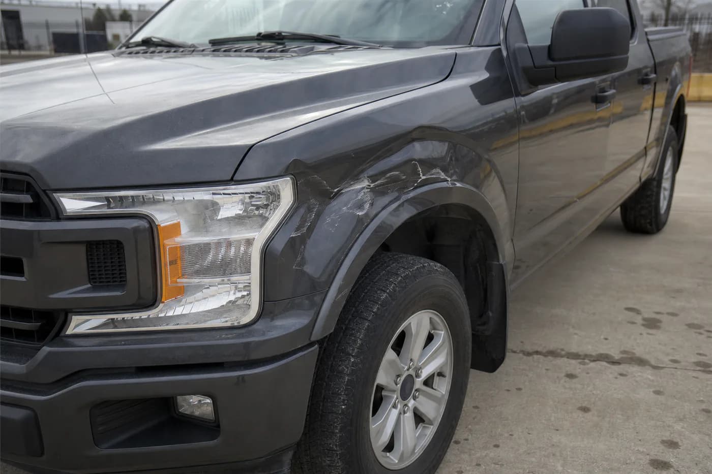 Dark gray pickup with a deep dent and paint damage on the left front fender